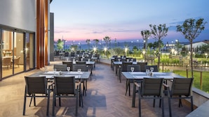 Un área de comedor al aire libre con mesas y sillas, con vistas panorámicas del océano y las colinas al atardecer.