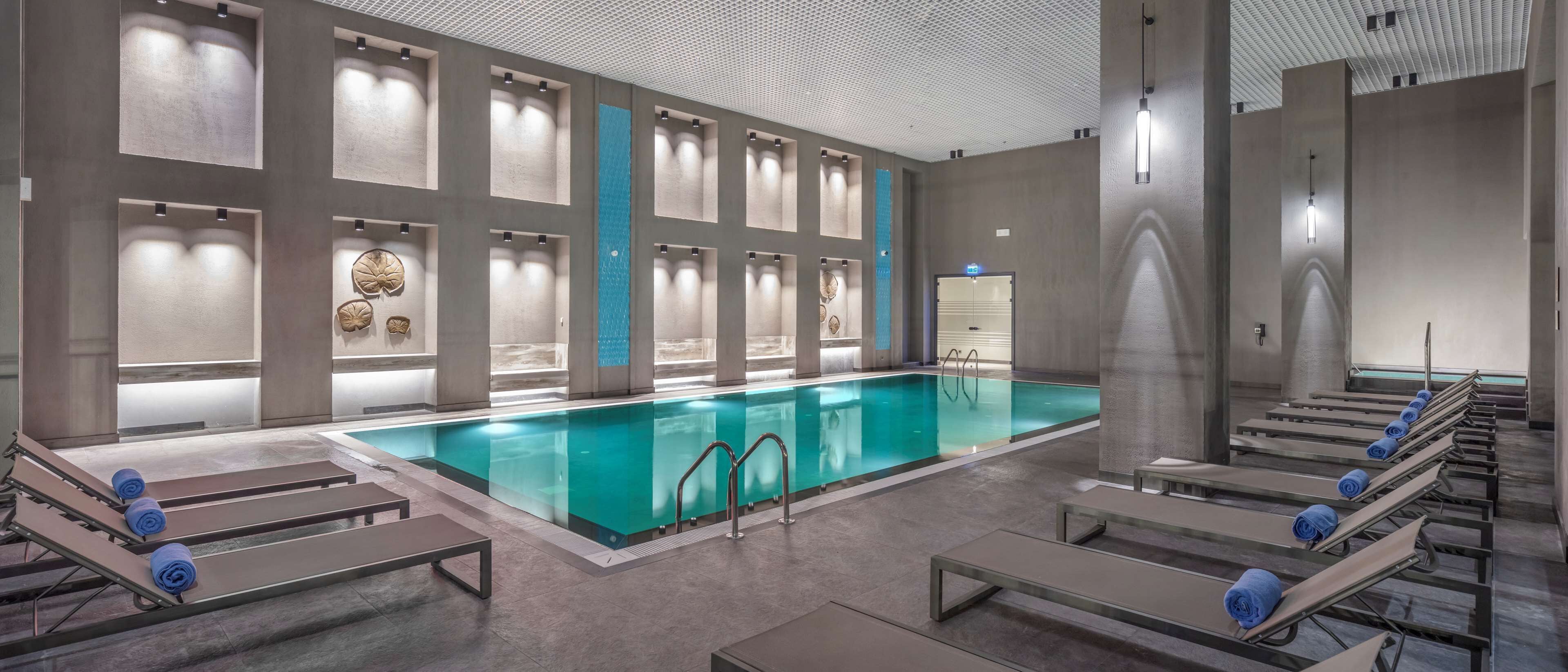A modern indoor swimming pool with clear water, surrounded by lounge chairs with towels, and a ceiling with a unique pattern.