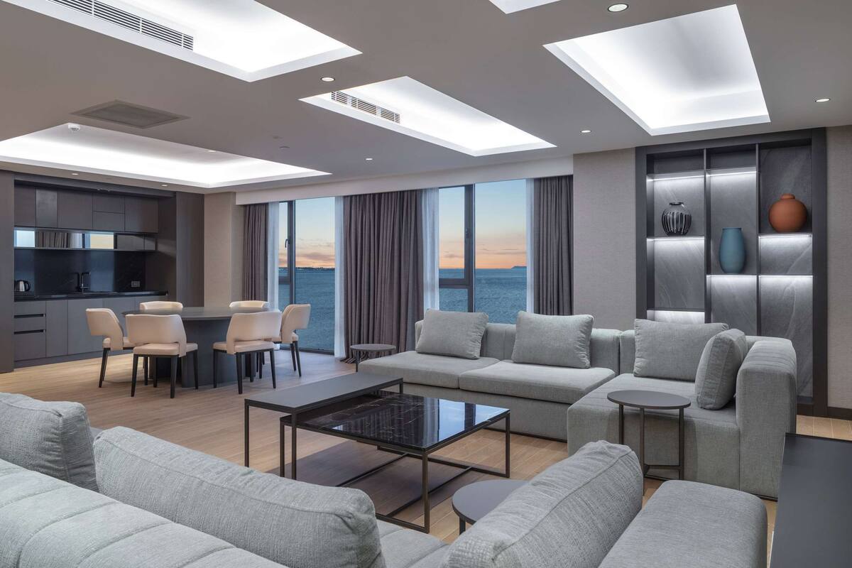 A modern living room with a sofa set, a coffee table, and a dining area with chairs. There is a large window offering a view of the sea.