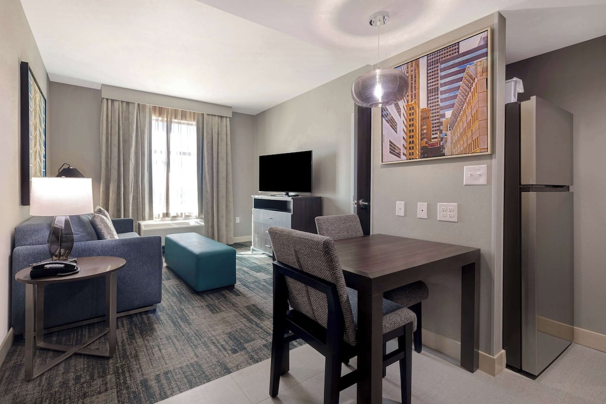 A modern hotel room with a living area, a dining table, a sofa, a TV, and a painting on the wall.