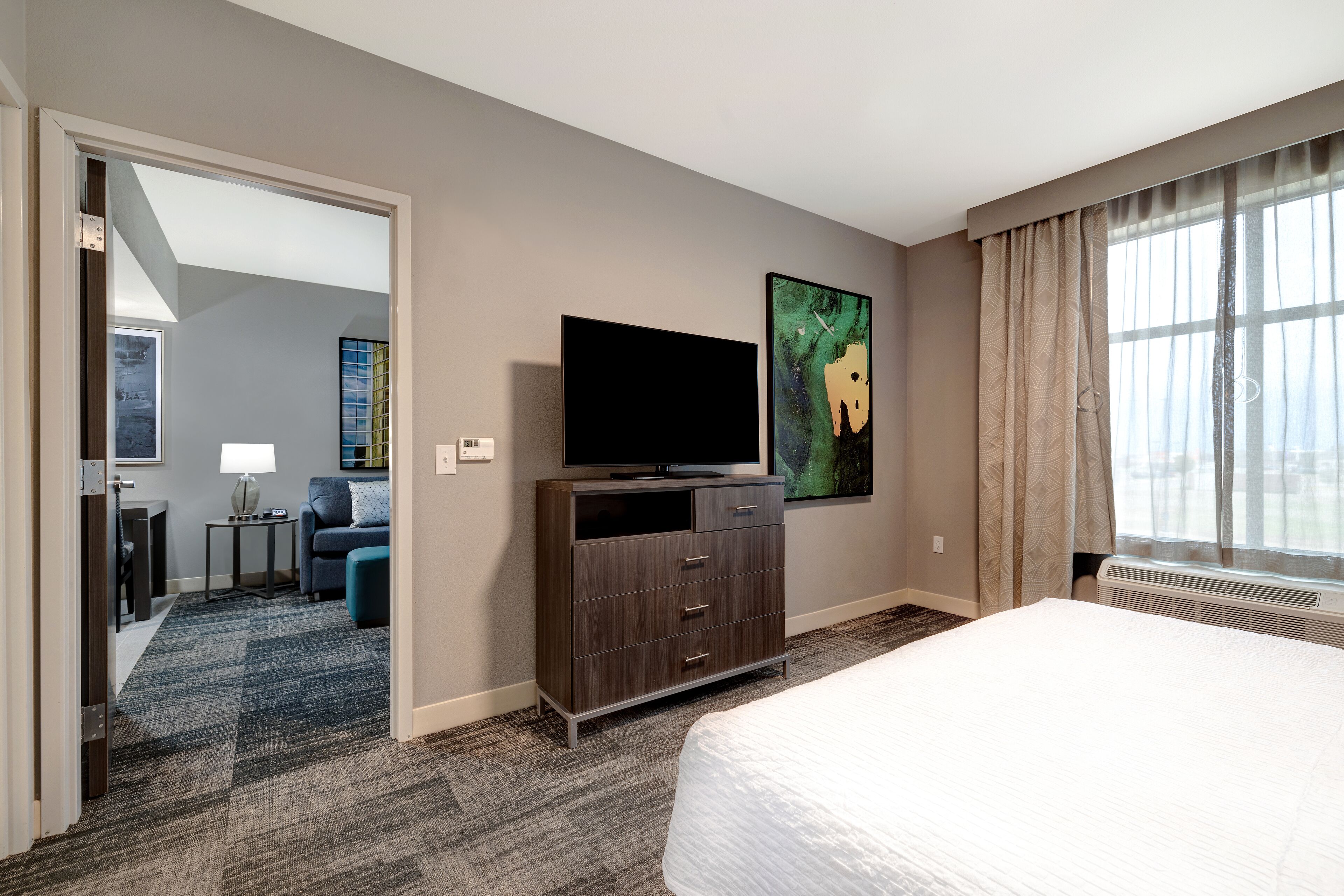 Habitación de hotel con una cama grande, un televisor de pantalla plana sobre un mueble de madera, un cuadro en la pared y una ventana con cortinas.