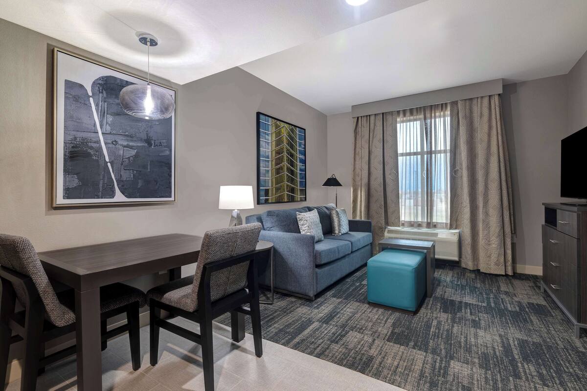 A modern hotel room with a sofa, a dining table with chairs, a blue ottoman, a TV, and a large window with curtains.