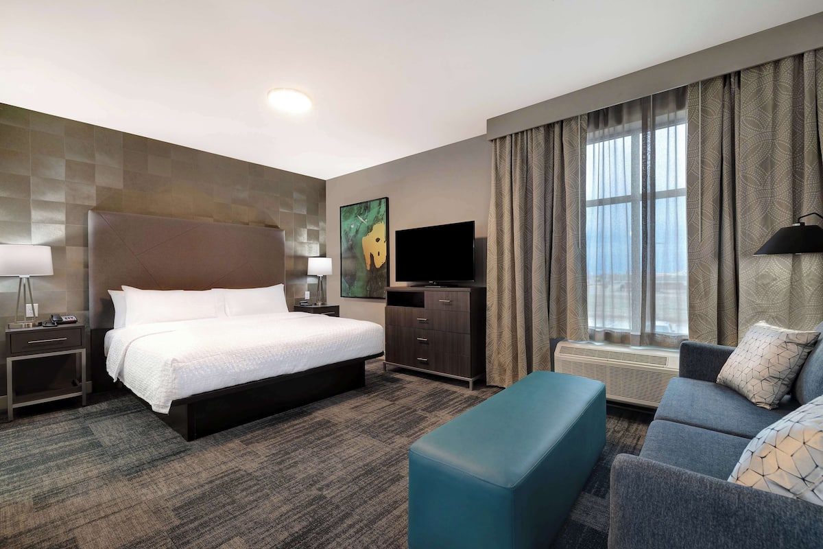 A hotel room with a large bed, a desk with a lamp, a television, a sofa, and a window with curtains.