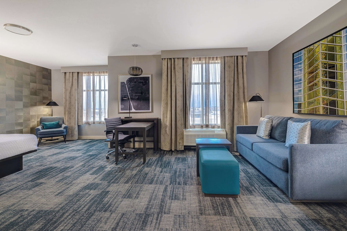 A hotel room with a bed, a desk, a sofa, and a large window with curtains.