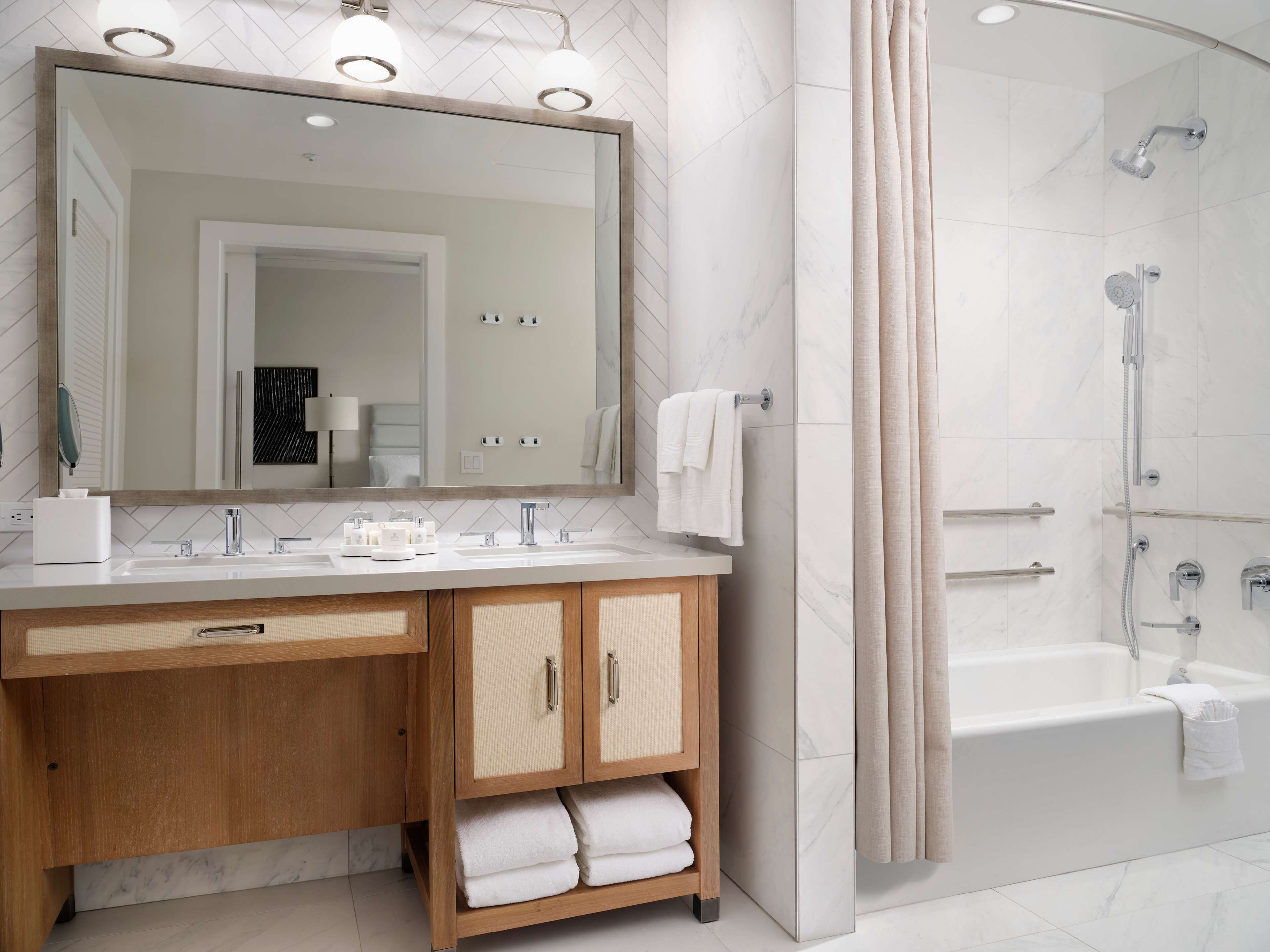 A modern bathroom with a large mirror, two sinks, wooden cabinets, and a bathtub.