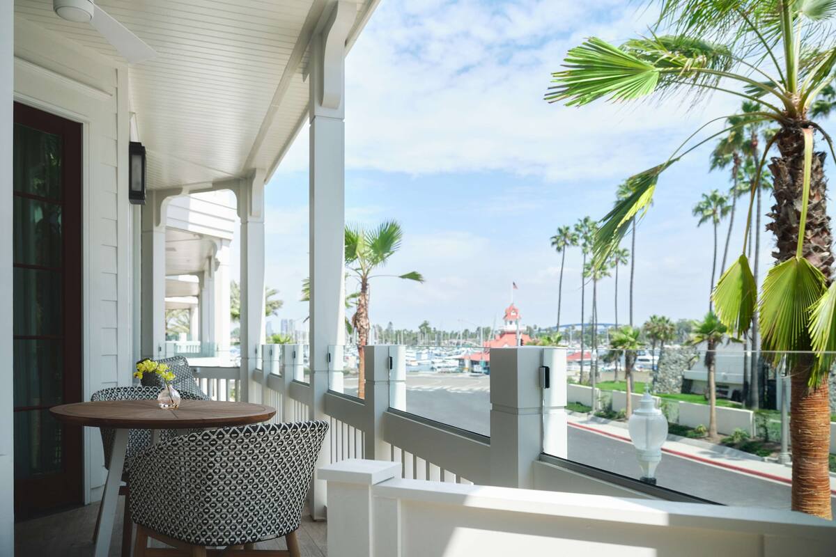 A covered balcony with a table and chairs, overlooking a street with palm trees and a red-roofed building.