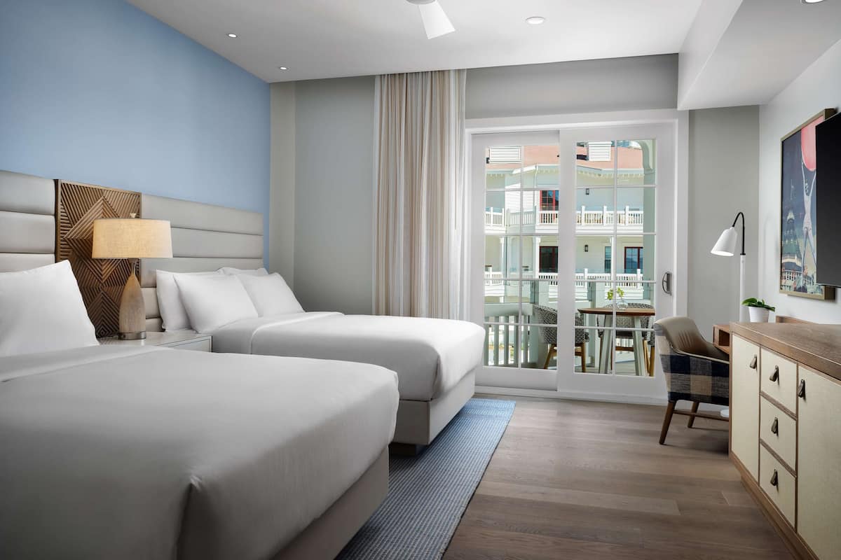 A hotel room with two beds, a desk, a chair, a ceiling fan, and a view of the outside balcony.