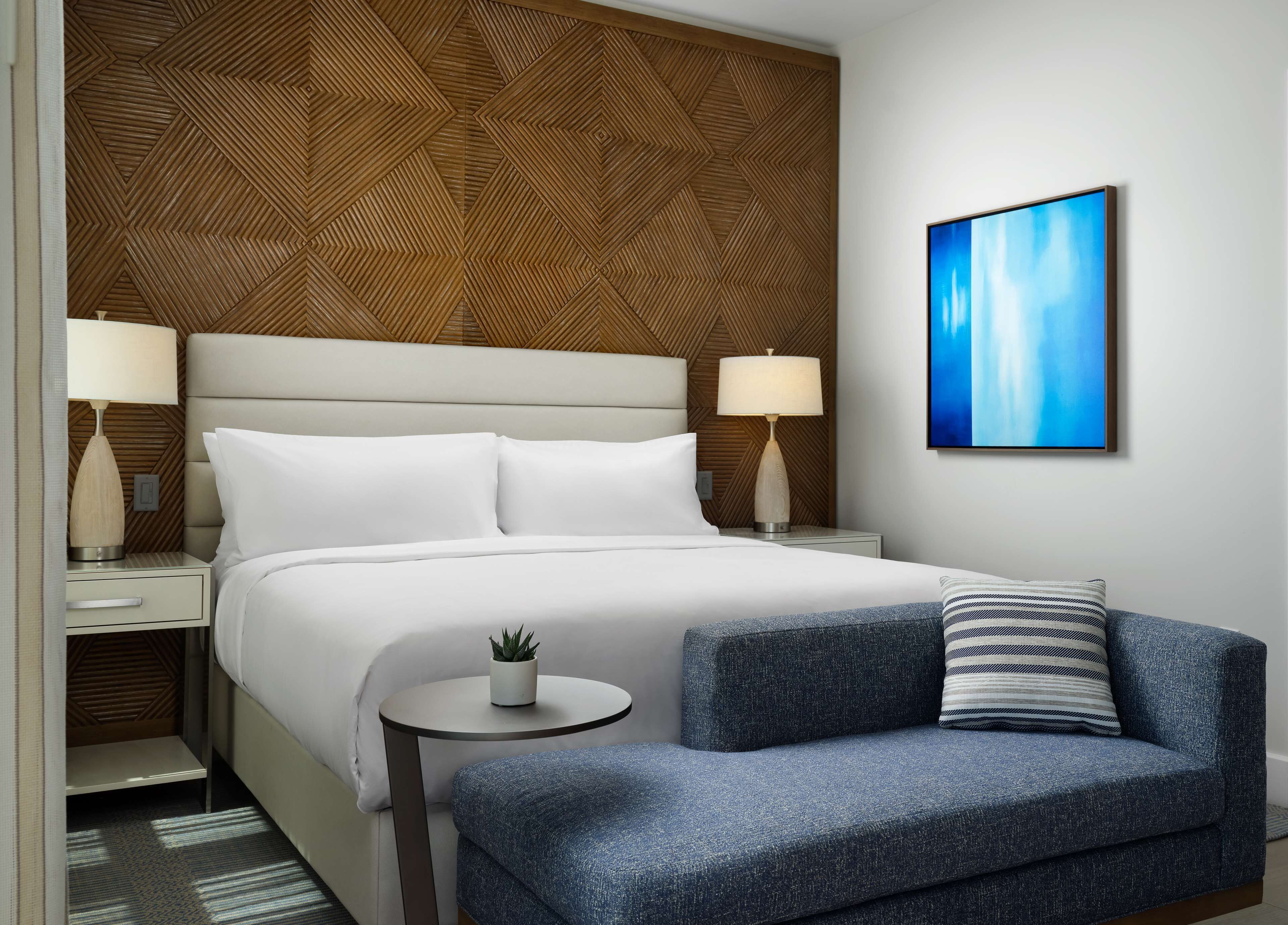 Ein modernes Hotelzimmer mit einem großen Bett, zwei Nachttischlampen, einem kleinen runden Tisch mit einer Pflanze, einem blauen Sessel und einem blauen abstrakten Gemälde an der Wand.