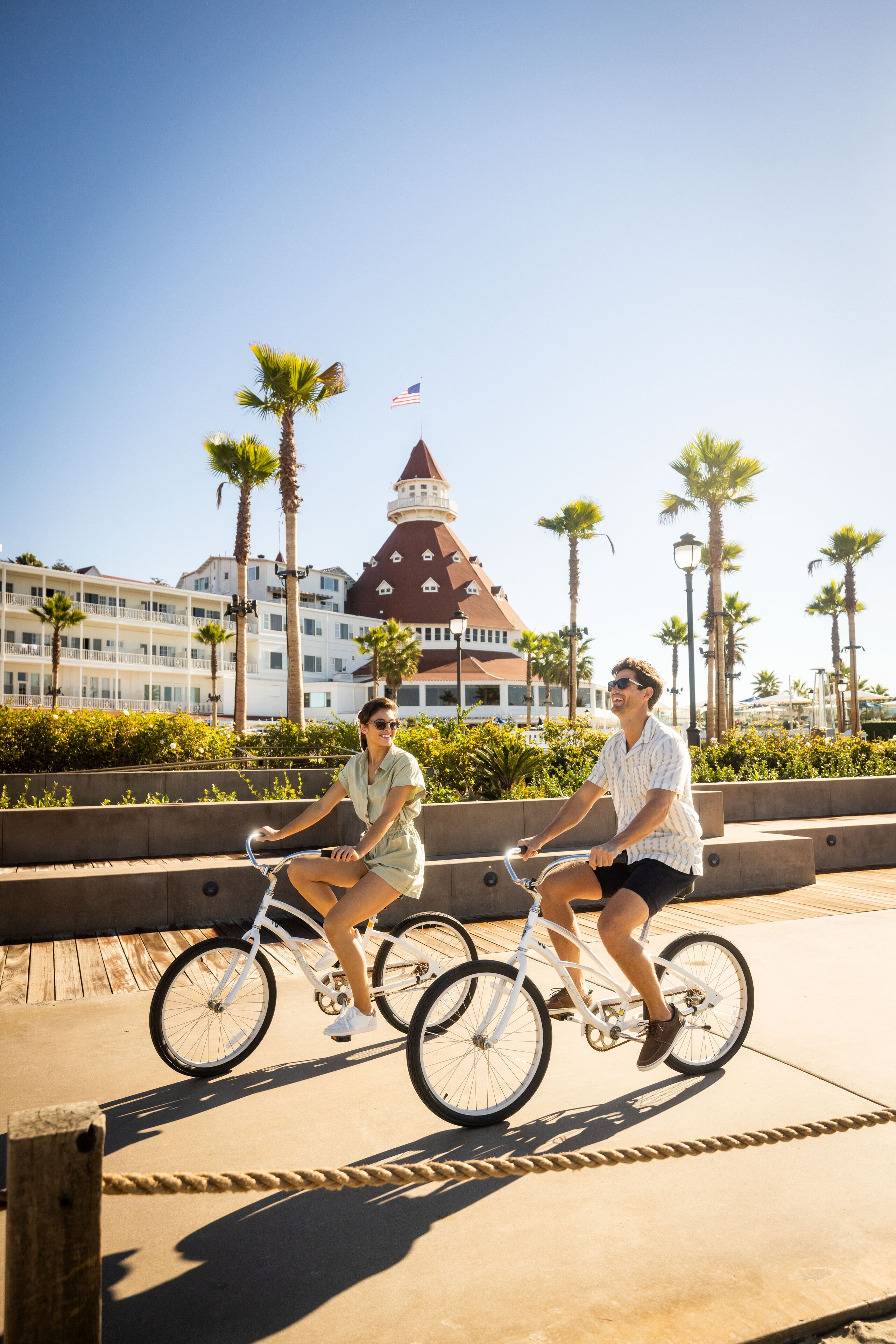 Two people riding bicycles on a paved path with palm trees and a building with a red roof in the background.