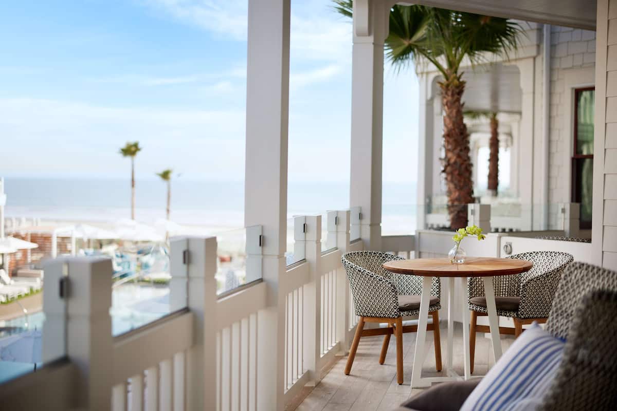 A balcony with a view of the beach, a round wooden table with two patterned chairs, and a palm tree.