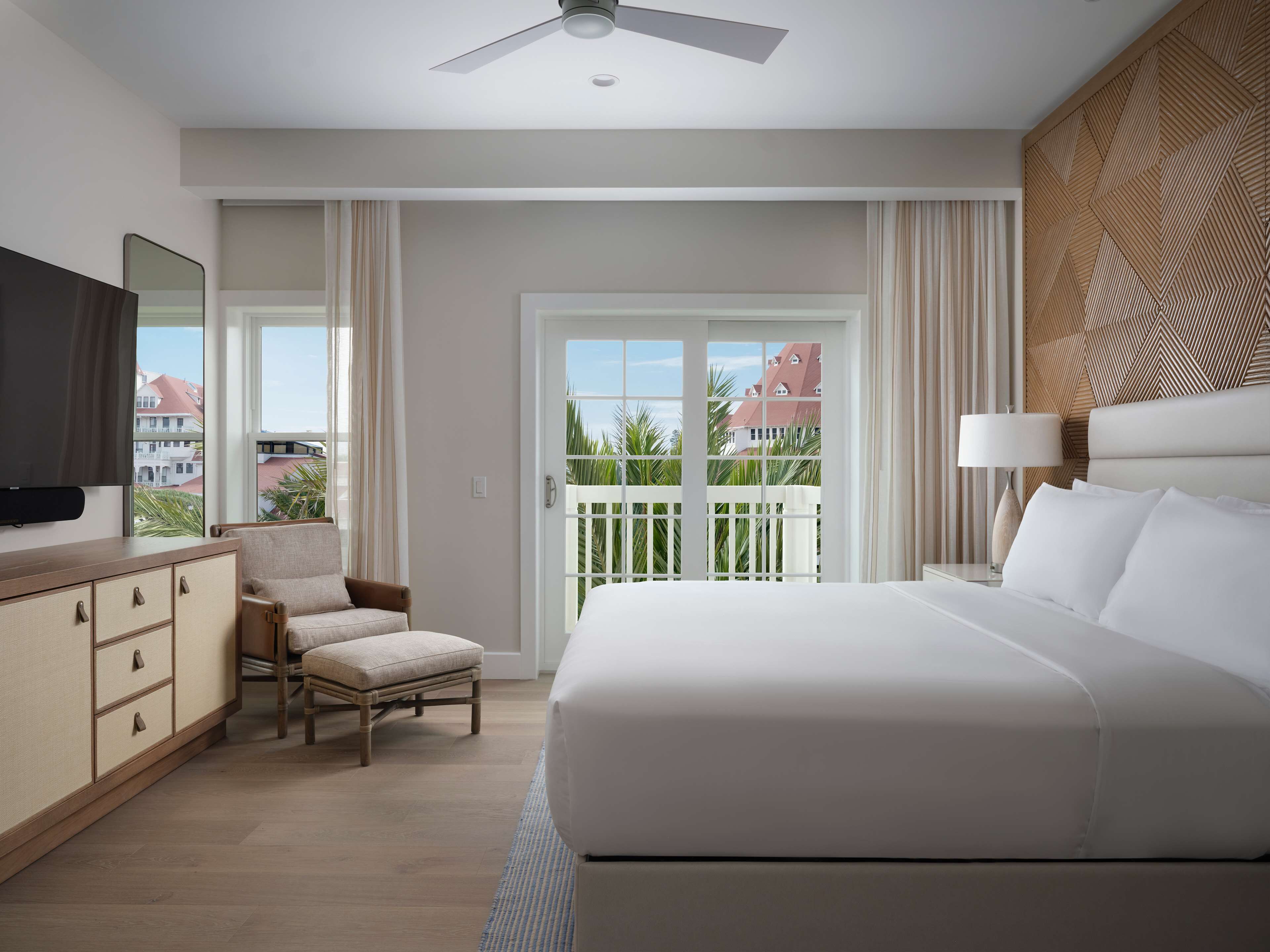 A hotel room with a large bed, a chair, a TV, a ceiling fan, and a balcony with a view of buildings and palm trees.