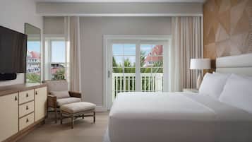 A hotel room with a large bed, a chair, a TV, a ceiling fan, and a balcony with a view of buildings and palm trees.