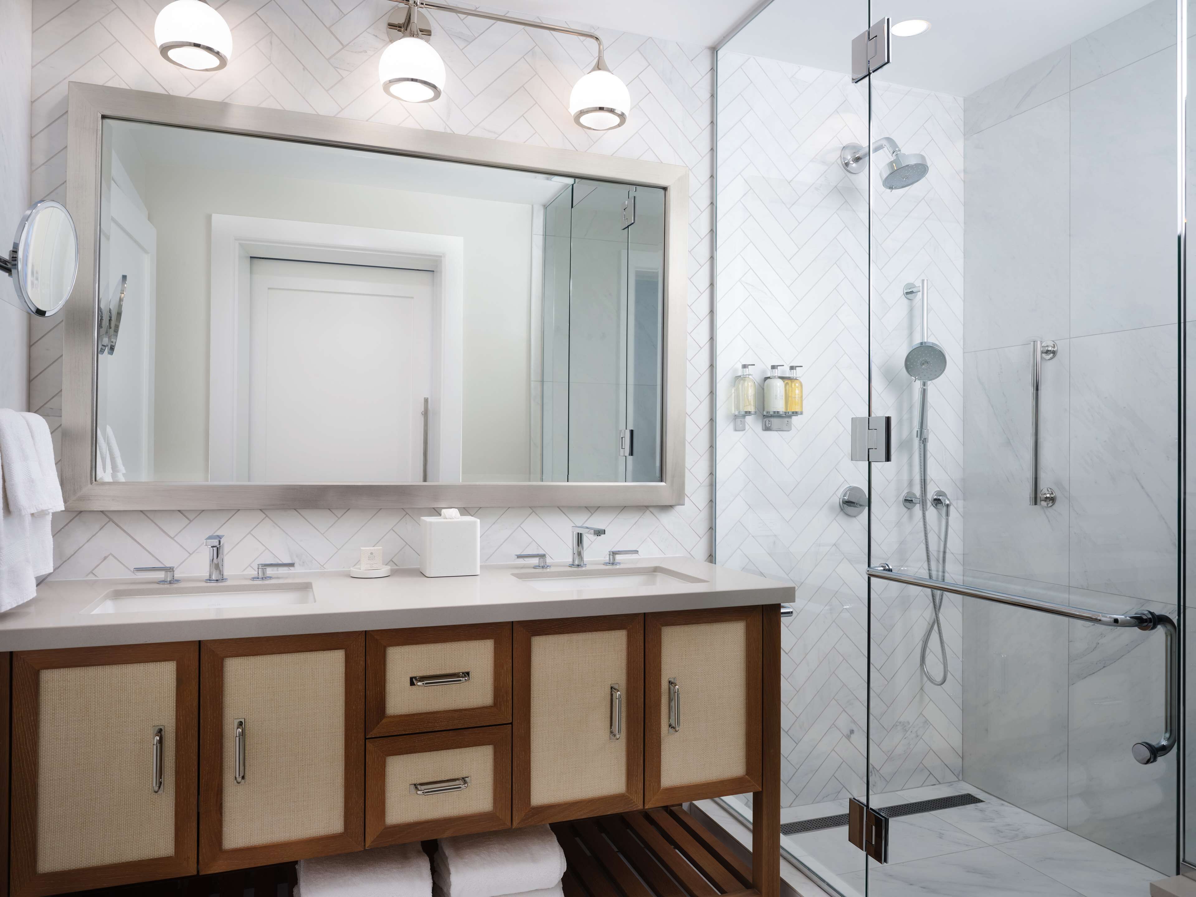 A modern bathroom with a large mirror, two sinks, wooden cabinets, and a glass-enclosed shower area.