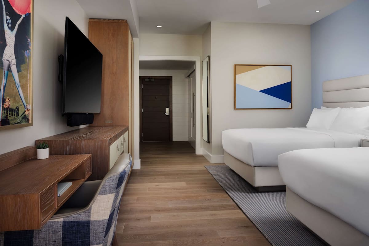 A modern hotel room with a flat-screen TV, a wooden console table, two beds, a ceiling fan, and a hallway leading to another room.