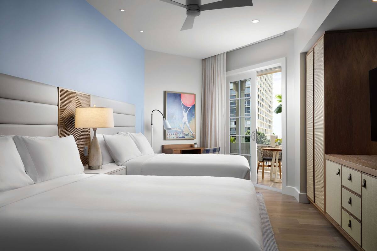 A modern hotel room with two beds, a ceiling fan, a desk with a chair, a TV, and a large window with a view of the city.