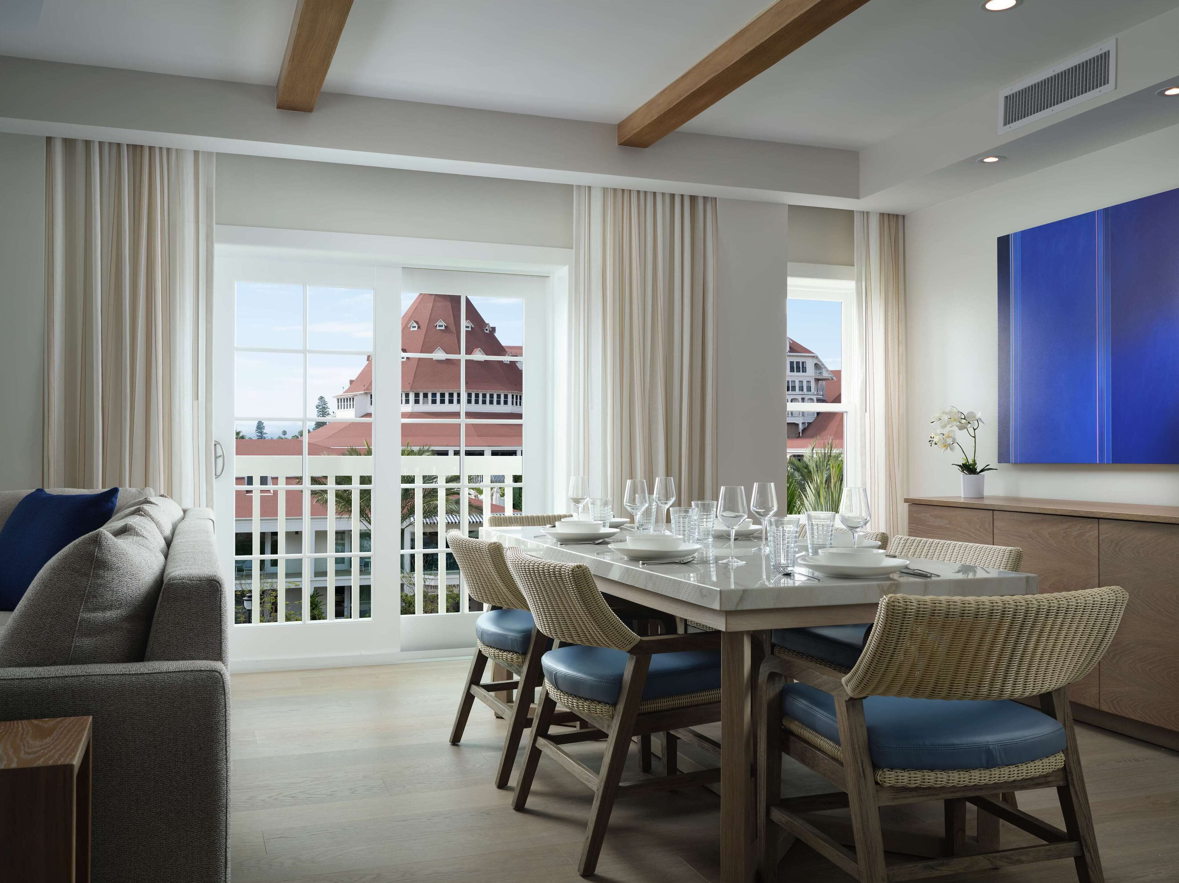A dining area with a table set for six, large windows, and a view of a building with a red roof.