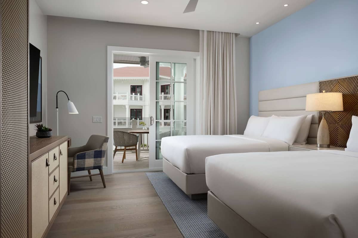 A hotel room with two beds, a desk, a chair, a TV, and a balcony with a view of the building.