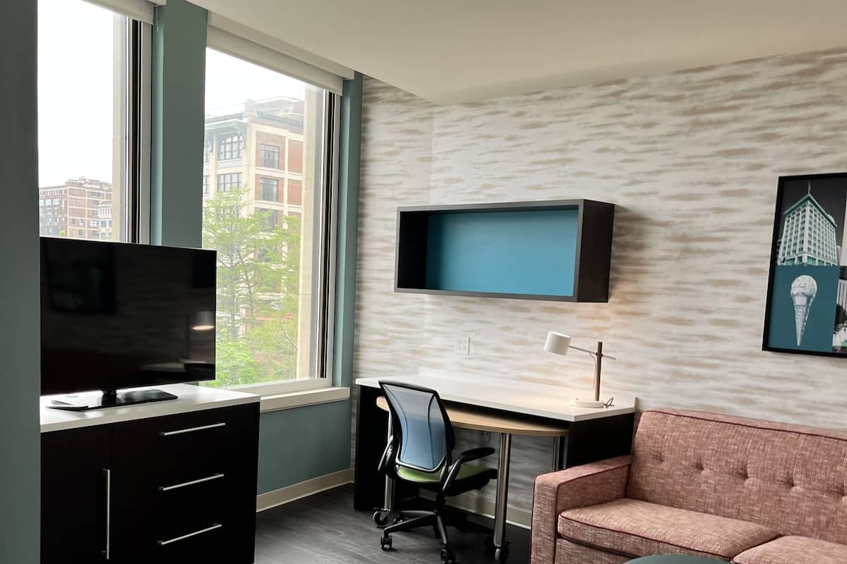 A modern living room with a desk, a sofa, a TV mounted on the wall, and a window with a view of buildings.