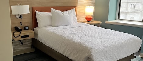 A hotel room with a large bed, a nightstand with a lamp, a wall-mounted phone, and a window with a view of buildings.