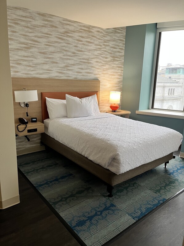 A hotel room with a large bed, a nightstand with a lamp, a wall-mounted phone, and a window with a view of buildings. - Home2 Suites by Hilton St. Louis Downtown (St. Louis)