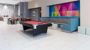 A pool table in a modern lounge area with a colorful geometric wall art and a gray sofa with patterned ottomans. - Home2 Suites by Hilton St. Louis Downtown (St. Louis)