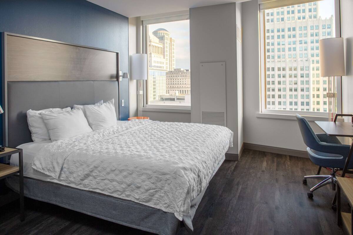 A modern hotel room with a large bed, a desk, and a view of a cityscape through the window.