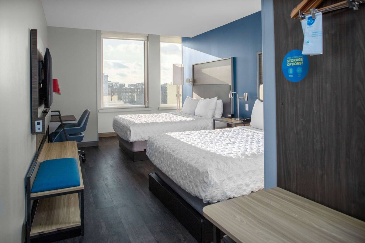 A hotel room with two beds, a desk, a chair, a TV, and a large window offering a city view.