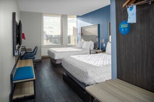 Habitación de hotel con dos camas, un escritorio, una silla, un televisor y un ventanal con vistas a la ciudad.