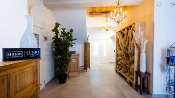 A hotel lobby with a wooden reception desk, a potted plant, a chandelier, and a wall decorated with wooden logs and pampas grass.