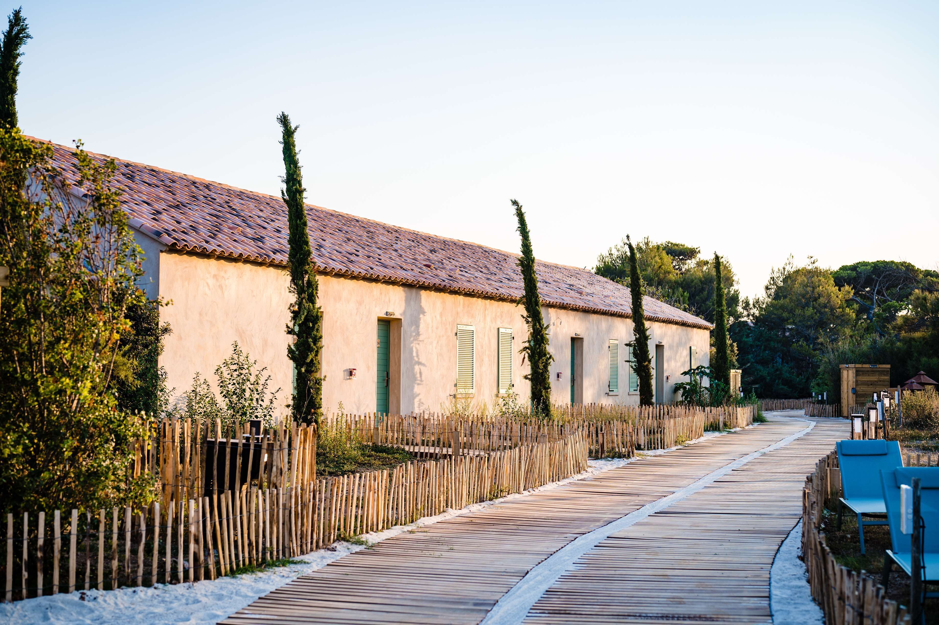 A wooden boardwalk leading to a row of buildings with a tiled roof, surrounded by greenery and a wooden fence.