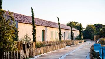 A wooden boardwalk leading to a row of buildings with a tiled roof, surrounded by greenery and a wooden fence.