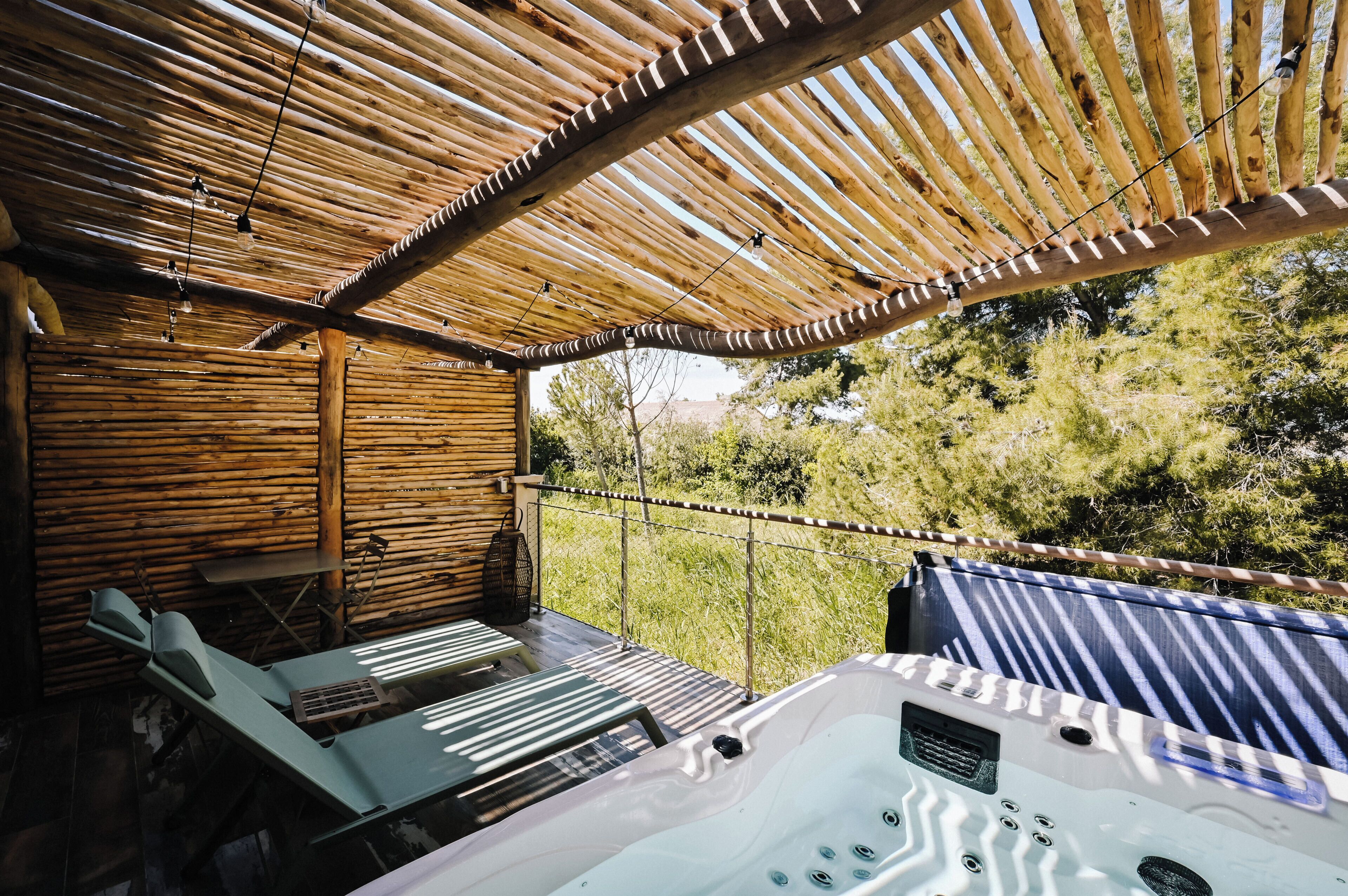 A wooden cabin with a hot tub, lounge chairs, and a view of the surrounding trees.