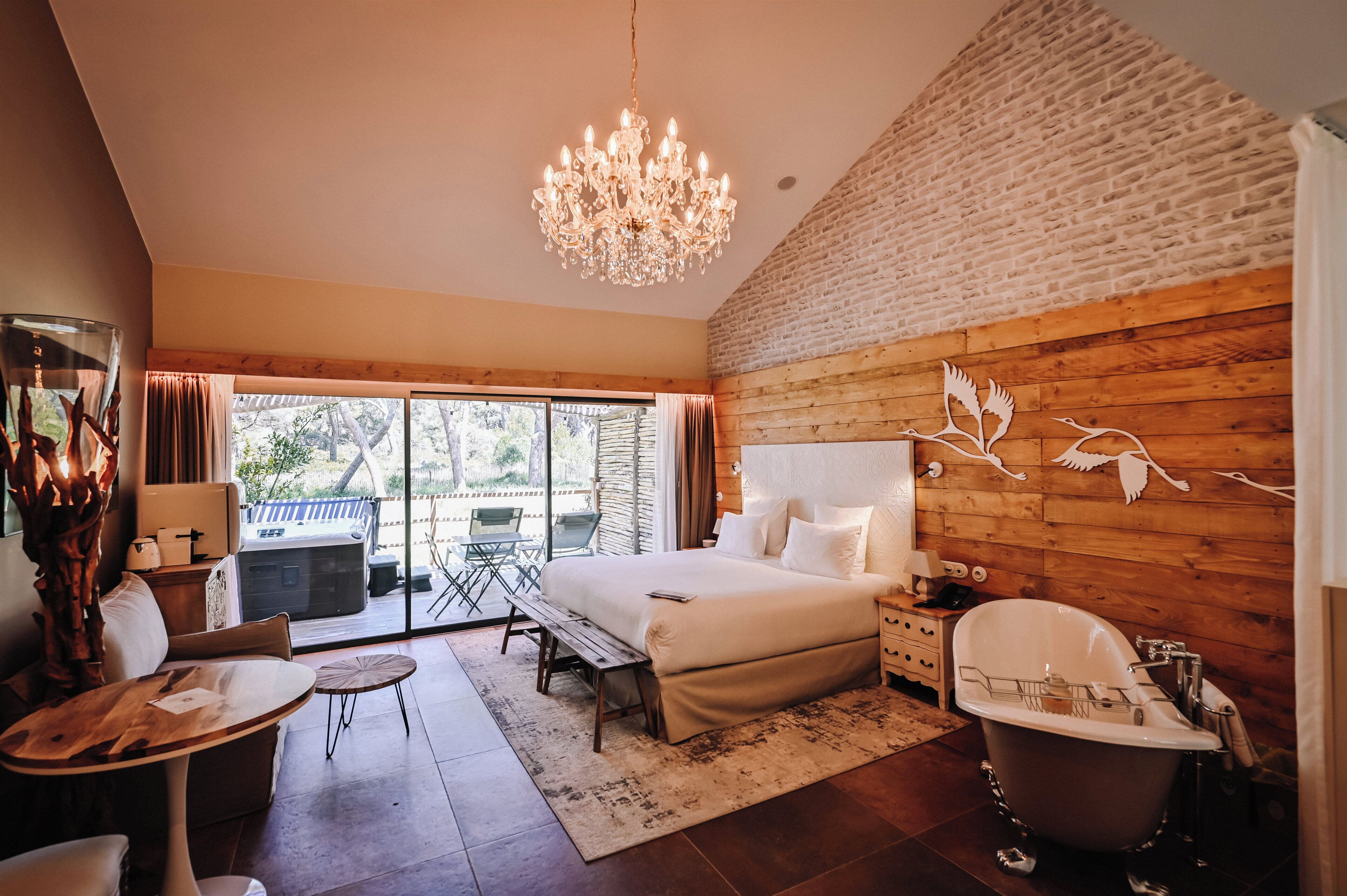A bedroom with a large bed, a chandelier, a wooden wall, and a view of the outdoors.