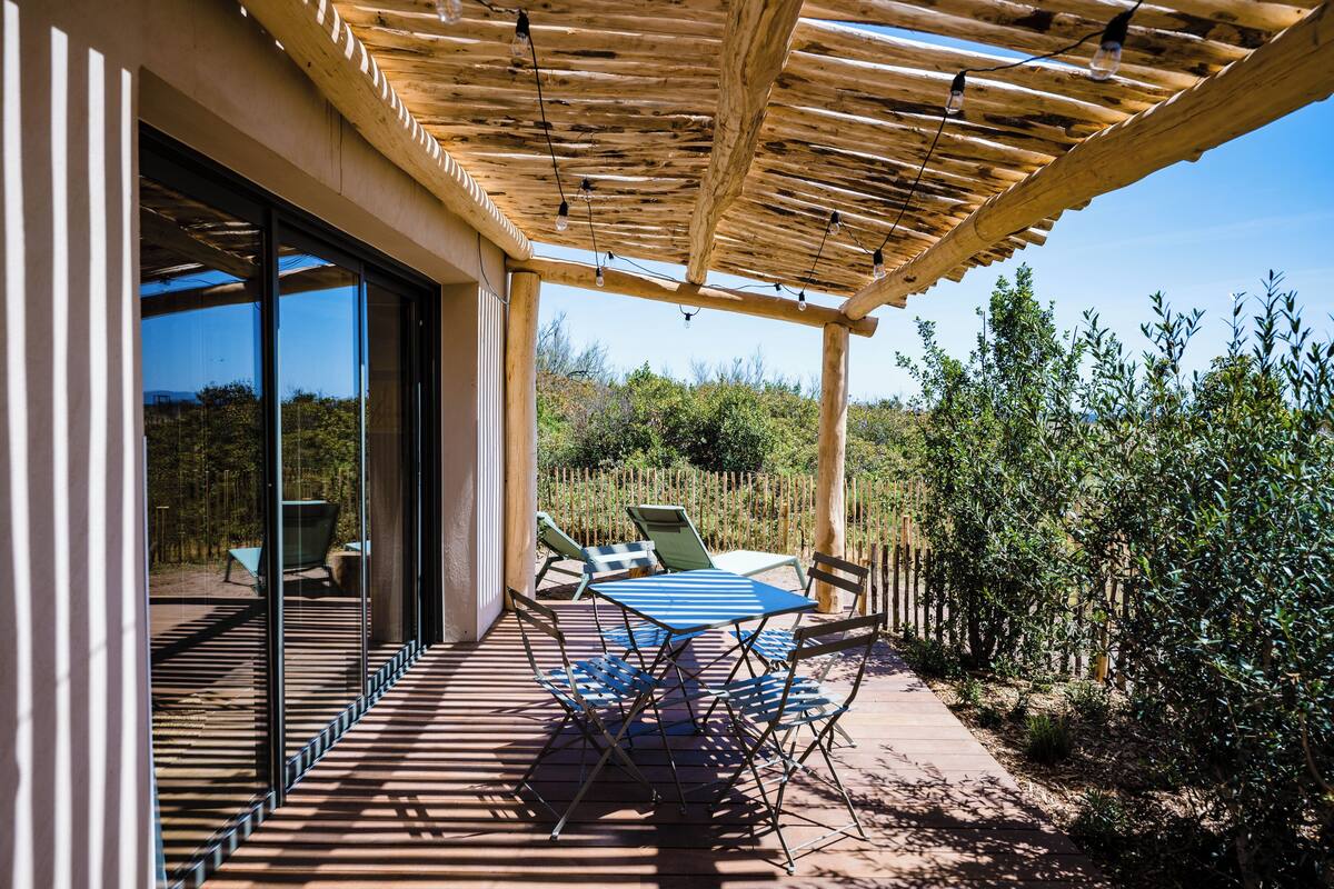 A covered outdoor patio with a table and chairs, a pergola with string lights, and a view of the surrounding vegetation.