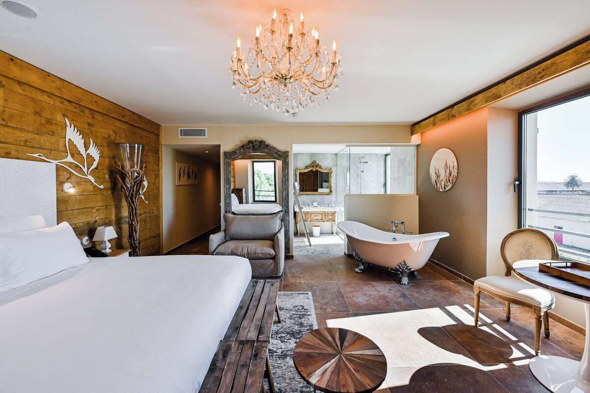 A modern hotel room with a large bed, a sitting area, a bathtub, and a view of the outside.