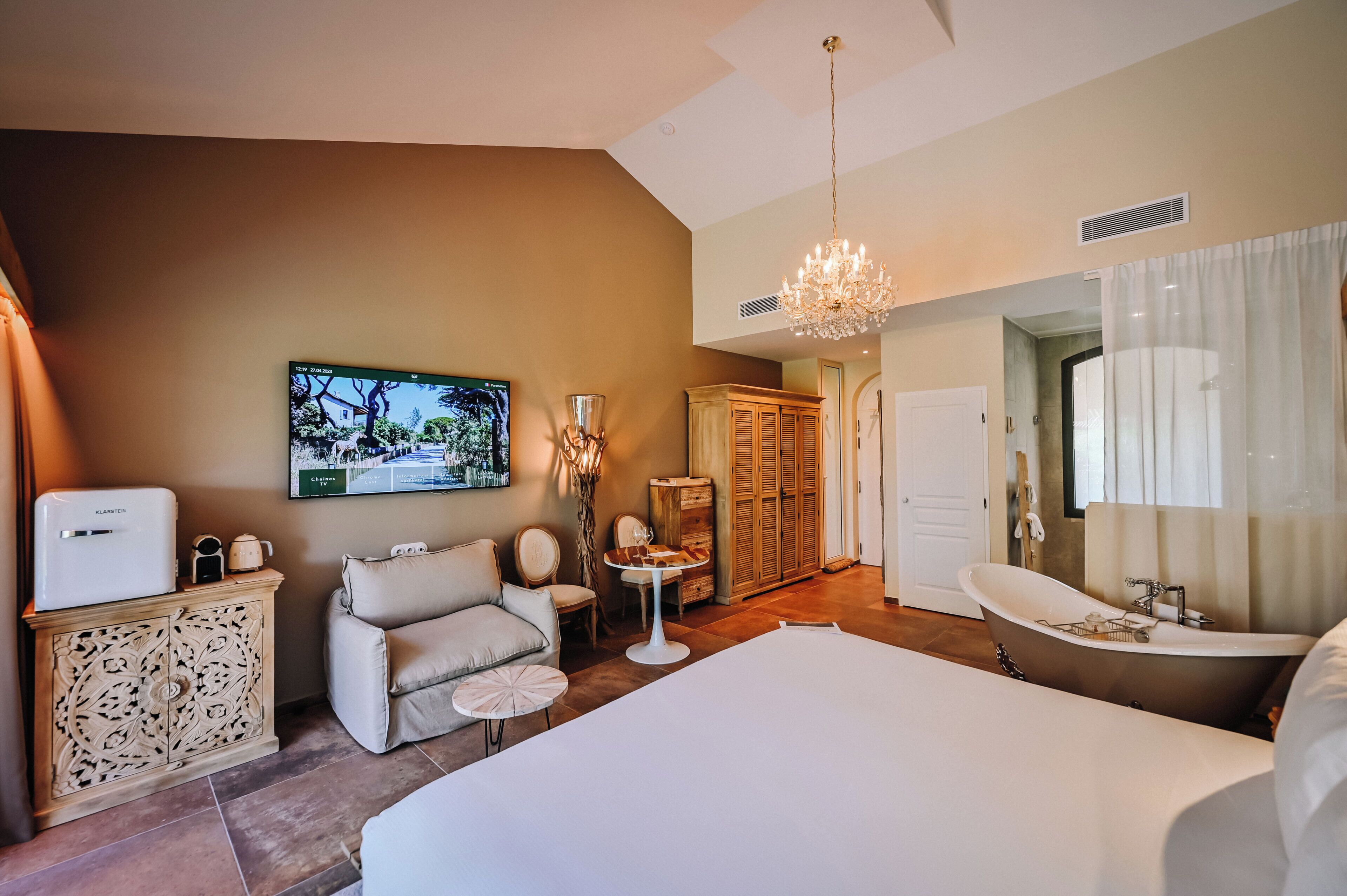 A bedroom with a large bed, a chandelier, a wooden cabinet, a chair, a small table, and a bathroom with a bathtub.