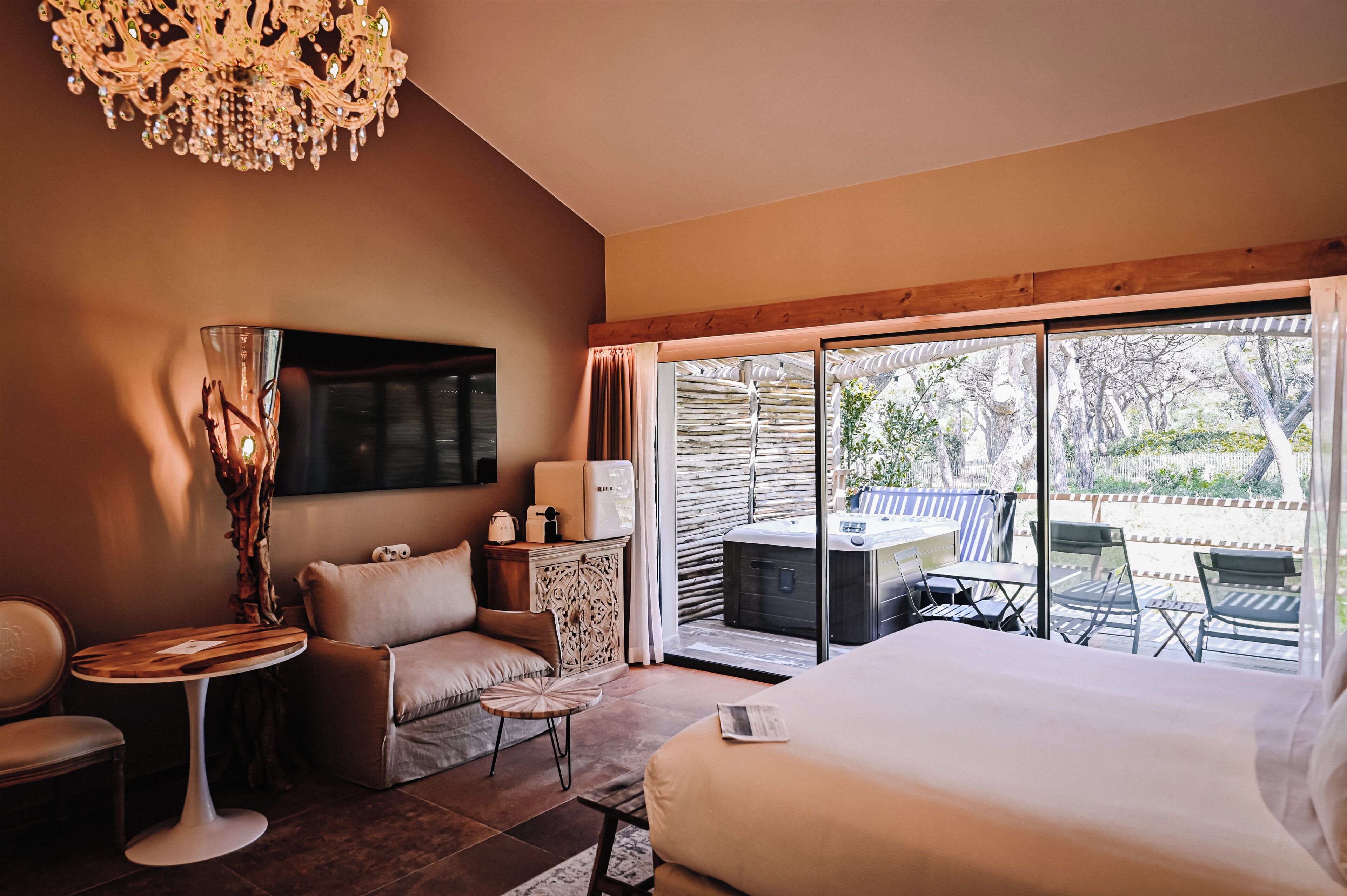 A hotel room with a bed, a sofa, a small table, a chandelier, a TV, and a sliding glass door leading to an outdoor area with a table and chairs.