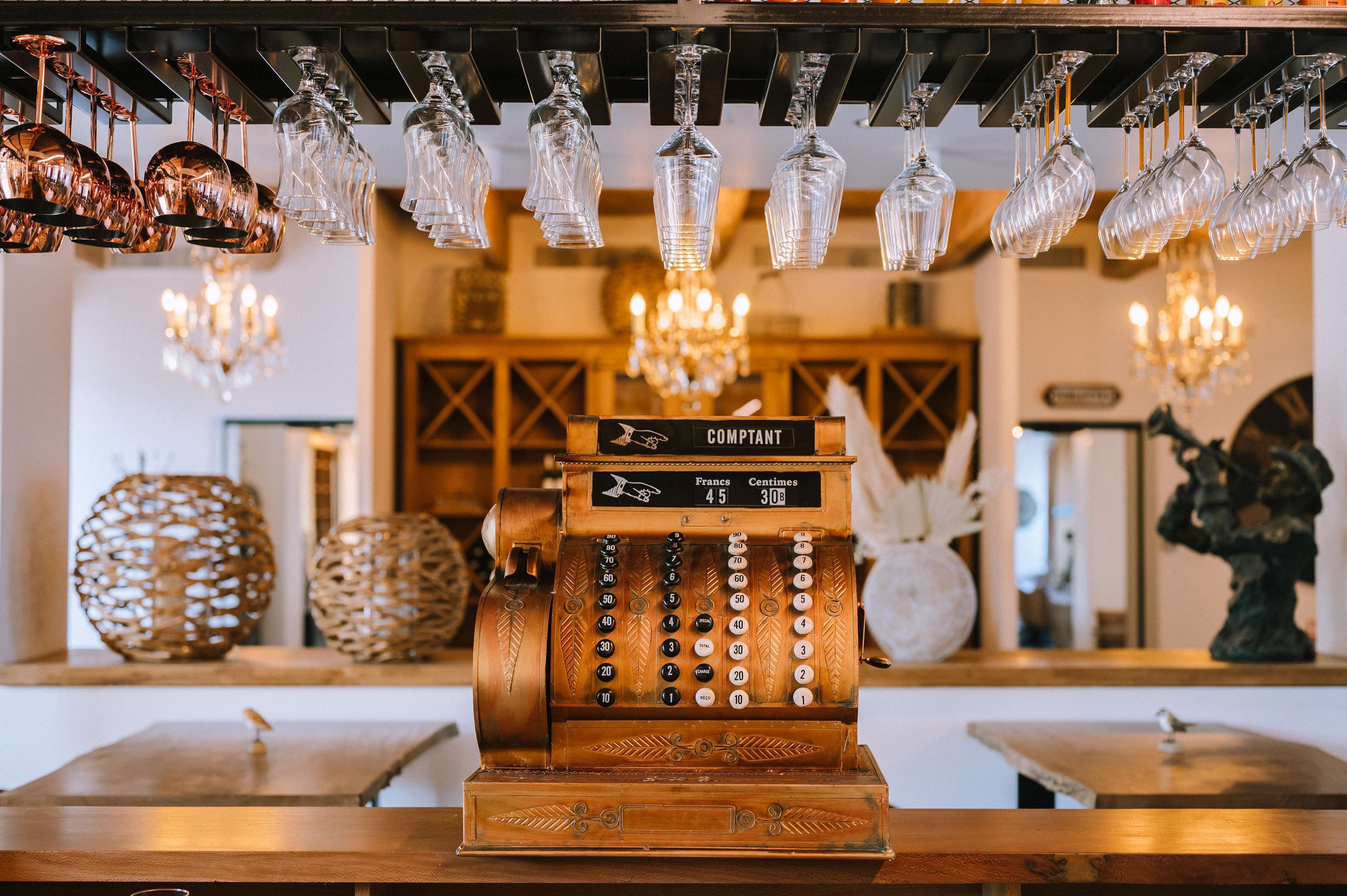 Une vieille caisse enregistreuse, accompagnée d’une liste de prix, entourée d’objets décoratifs et de verres suspendus.