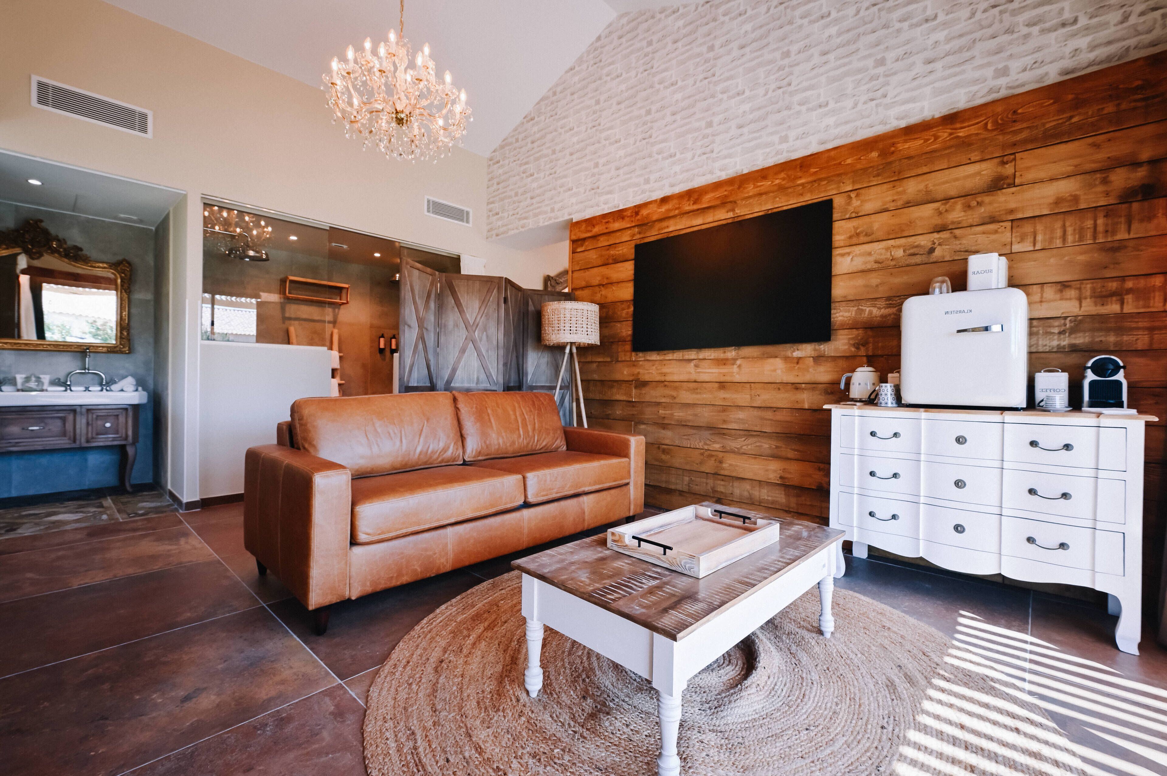 A modern living room with a brown leather sofa, a white coffee table, a wooden cabinet with a flat-screen TV, and a chandelier.