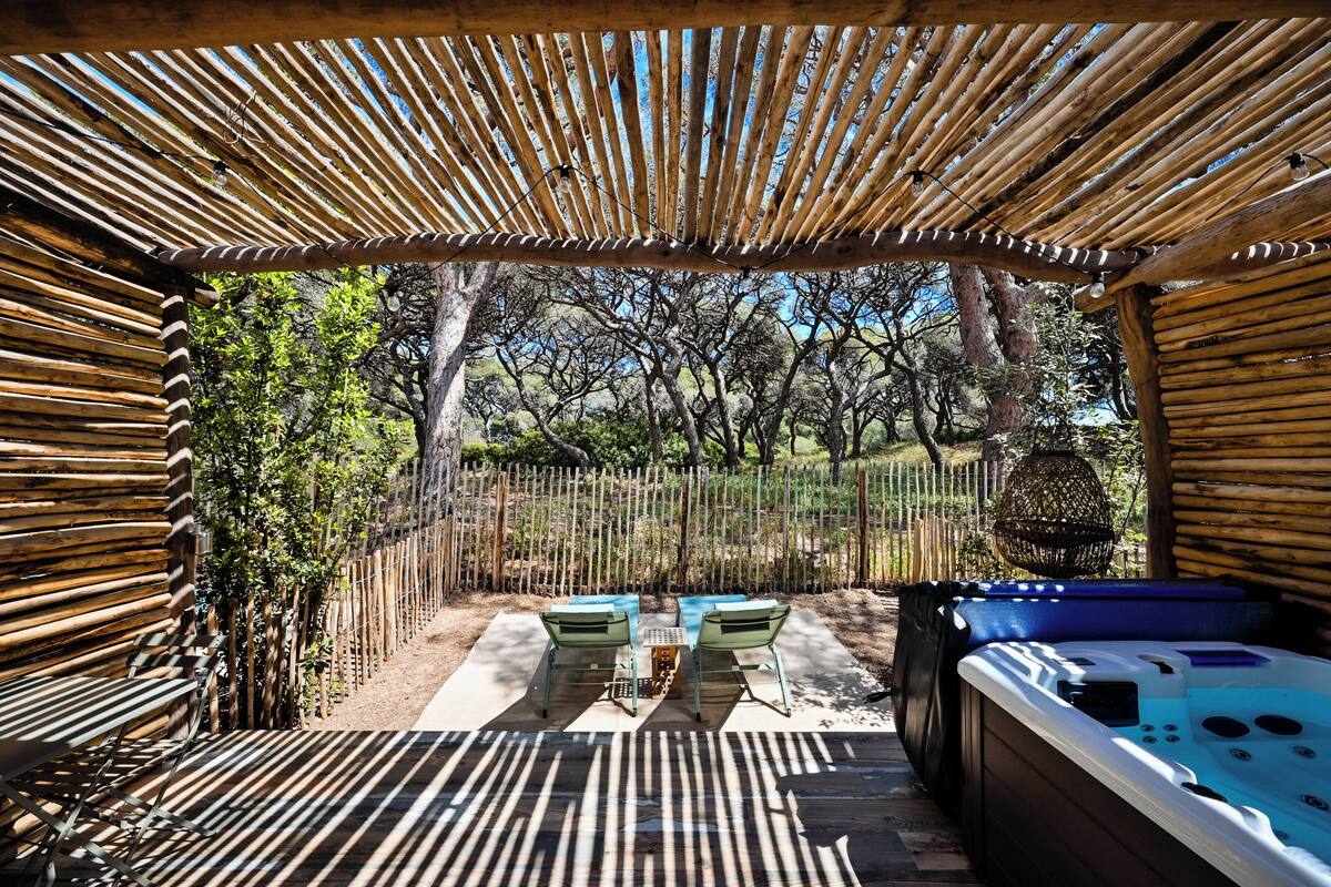 A covered outdoor seating area with a hot tub, wooden slatted roof, and a view of trees.