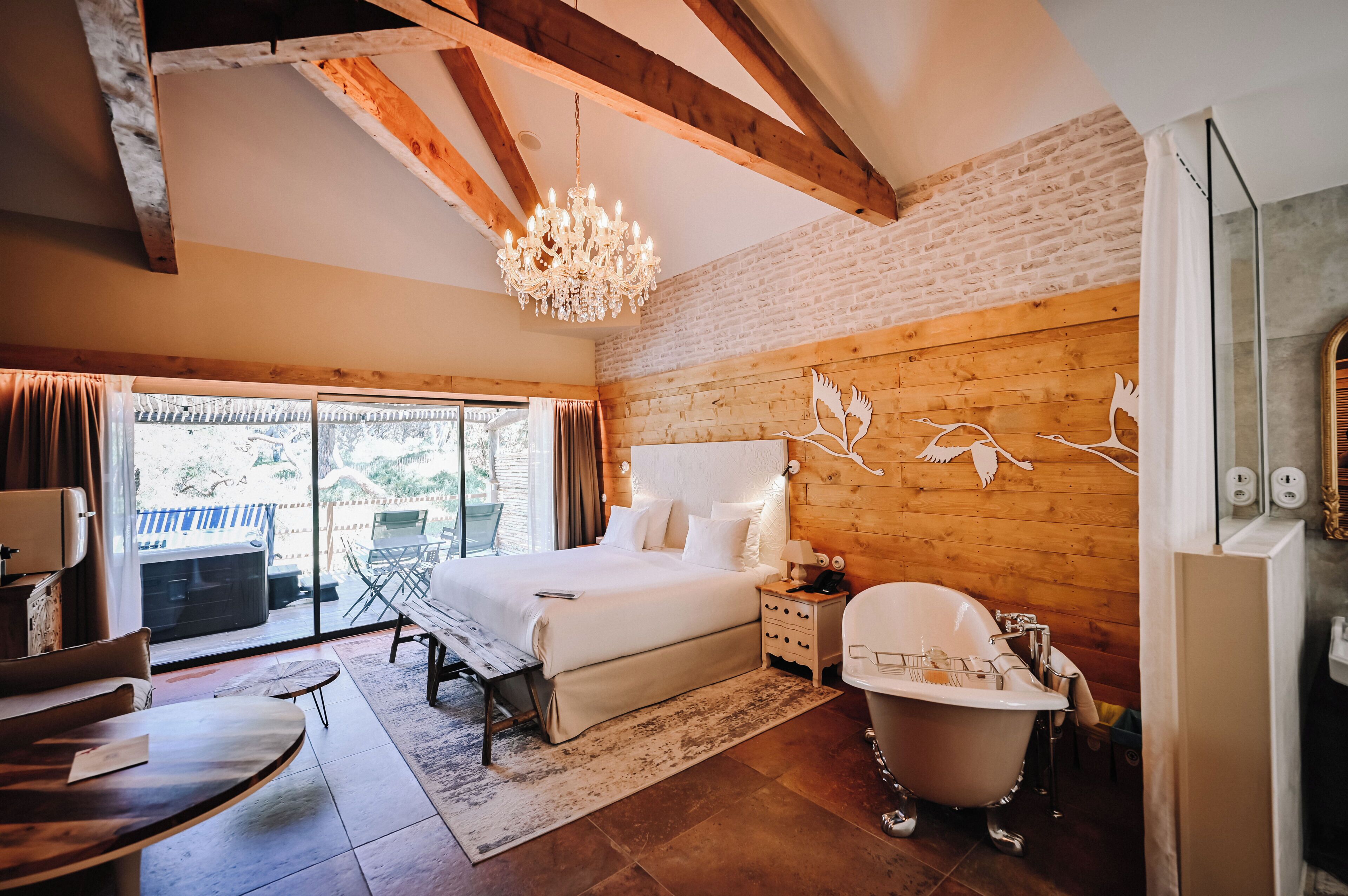 A bedroom with a large bed, a wooden ceiling, a chandelier, a bathtub, and a view through the window.