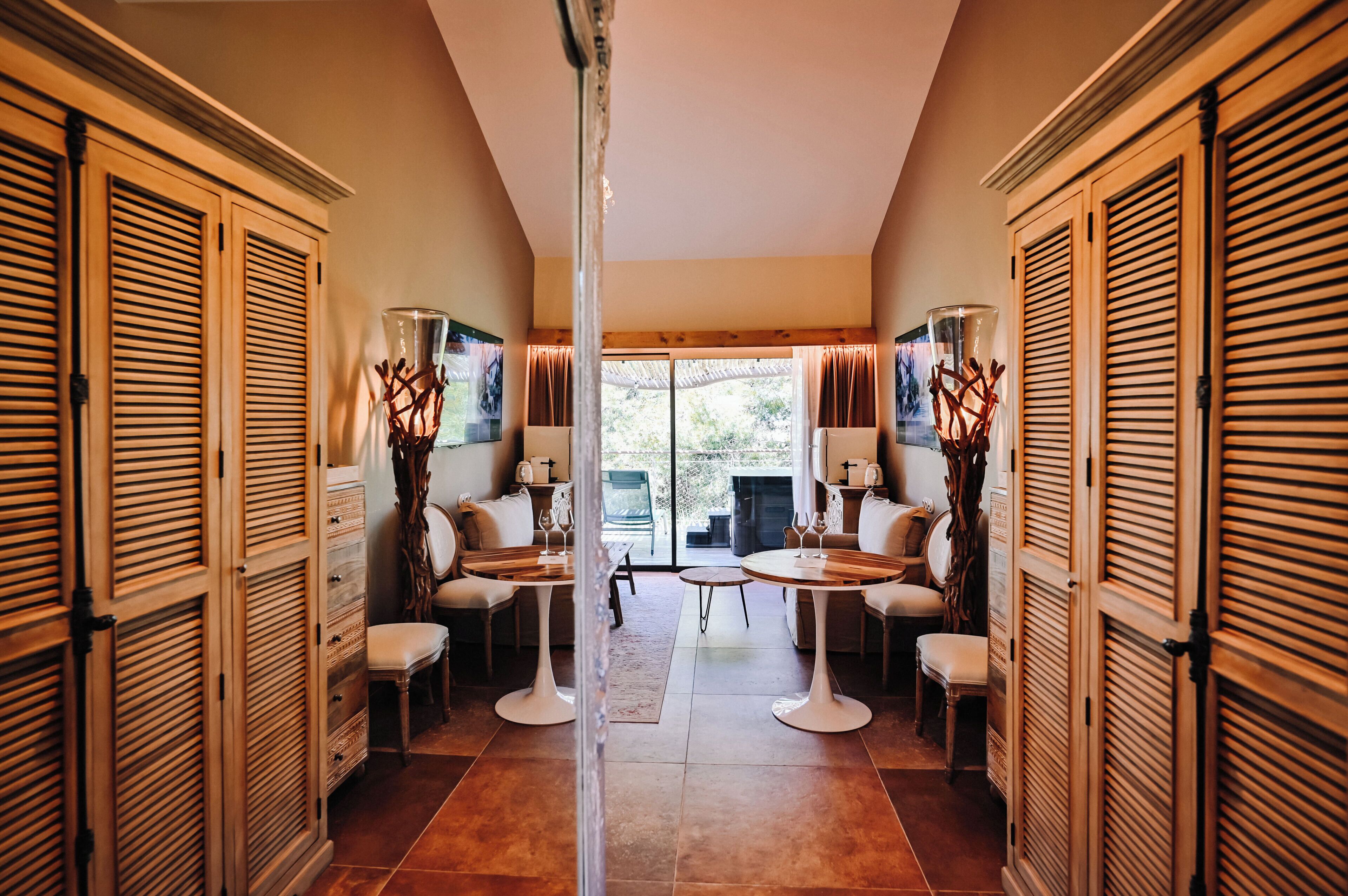 A hallway with wooden shutters, two round tables with chairs, and a view to the outside.
