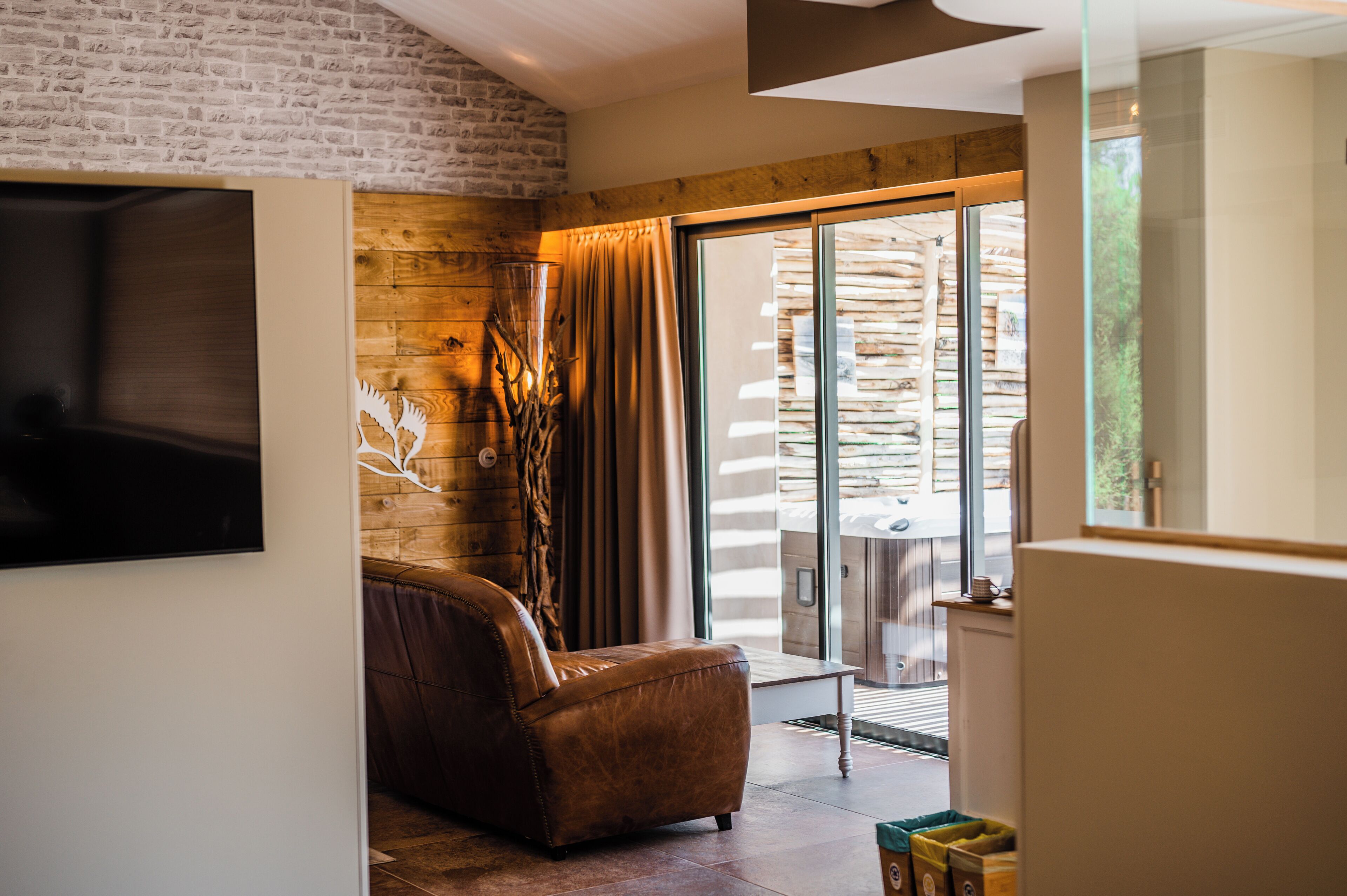 A living room with a leather sofa, a flat-screen TV mounted on the wall, and a sliding glass door leading to an outdoor area with a wooden deck.