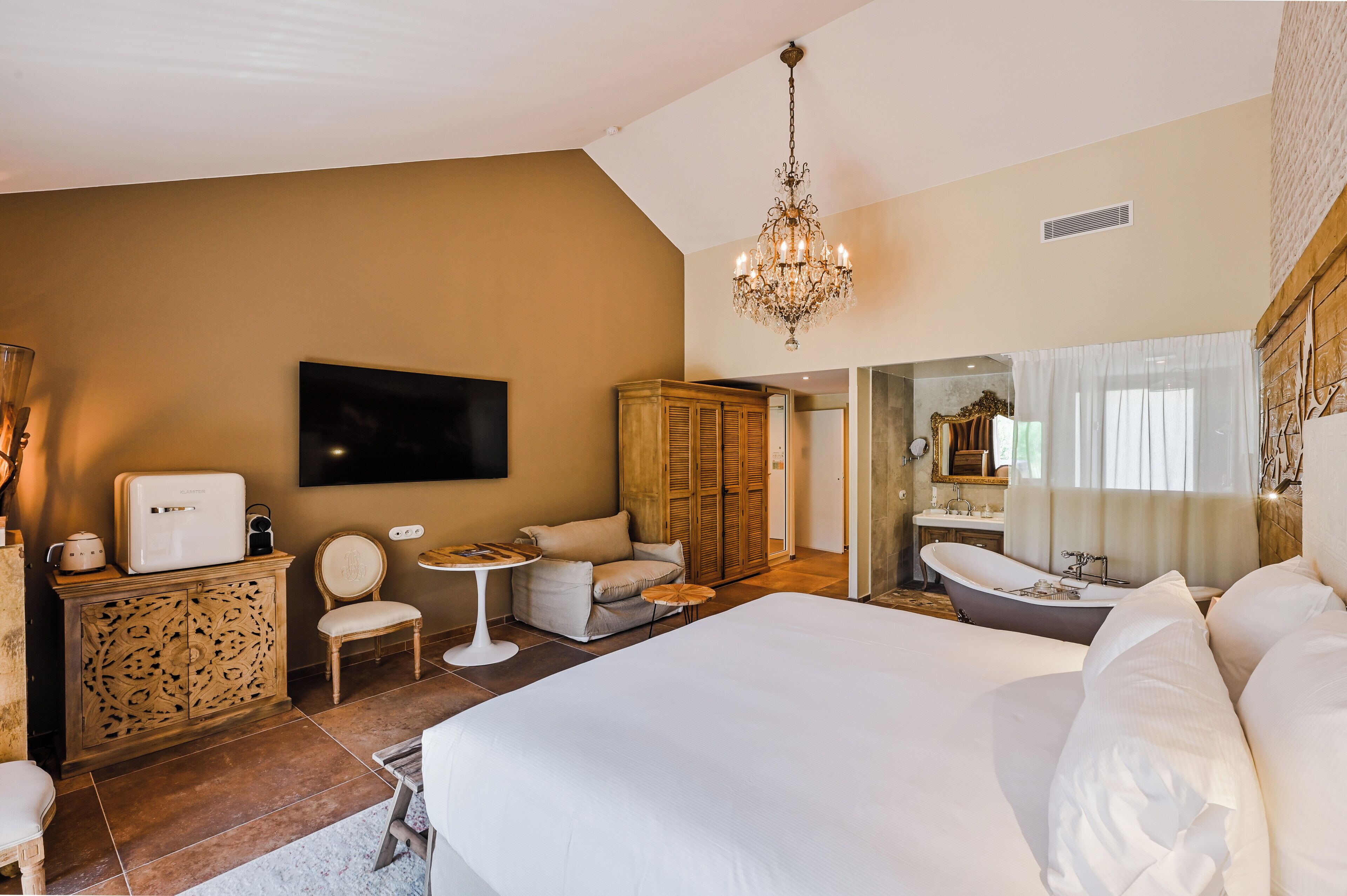A hotel room with a large bed, a sitting area with a table and chairs, a wardrobe, a television mounted on the wall, and a bathroom with a bathtub and mirror.