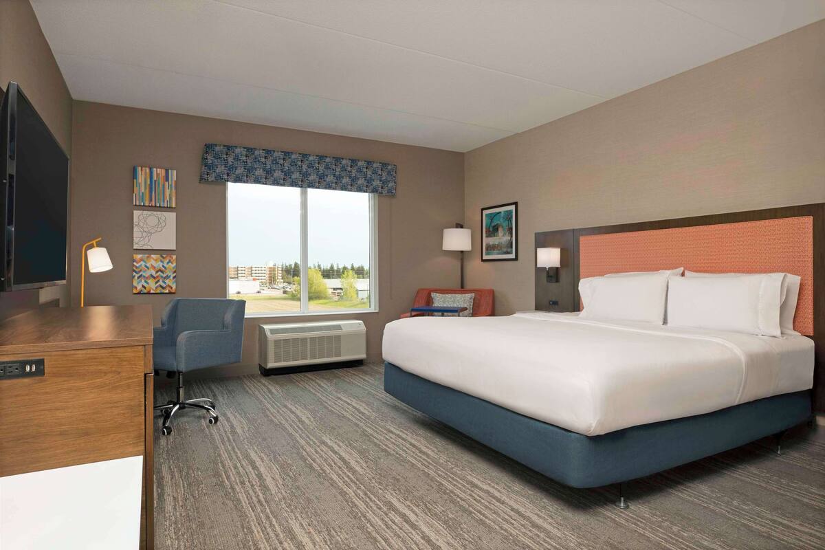A hotel room with a large bed, a desk, a chair, a window with a view, and a wall with artwork.