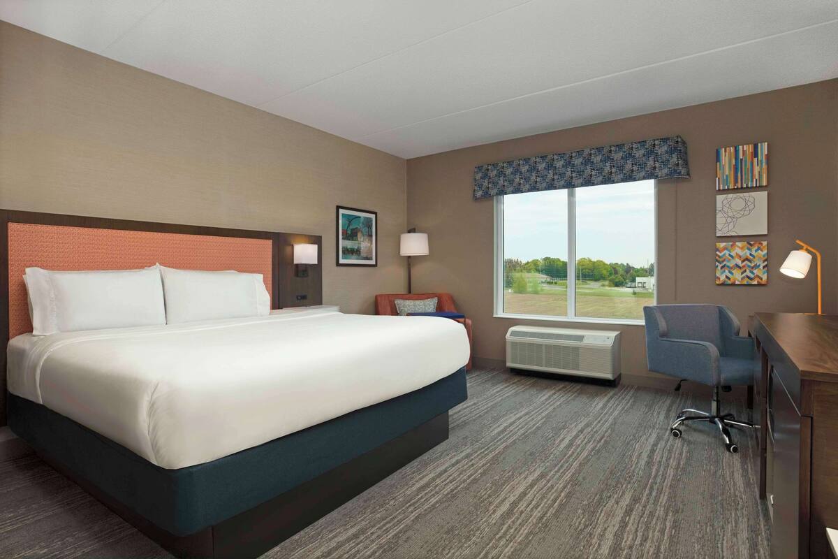 A hotel room with a large bed, a desk, a chair, a window with a view, and a wall with artwork.