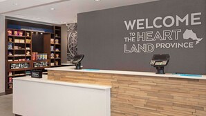 A hotel reception area with a welcome sign, a counter with a payment terminal, and a shelf with various snacks and toiletries. - Hampton Inn by Hilton Port Hope Cobourg (Port Hope)