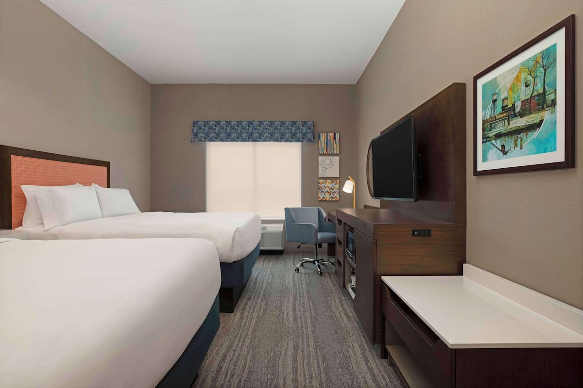 A hotel room with two beds, a desk, a chair, a TV, and artwork on the walls.