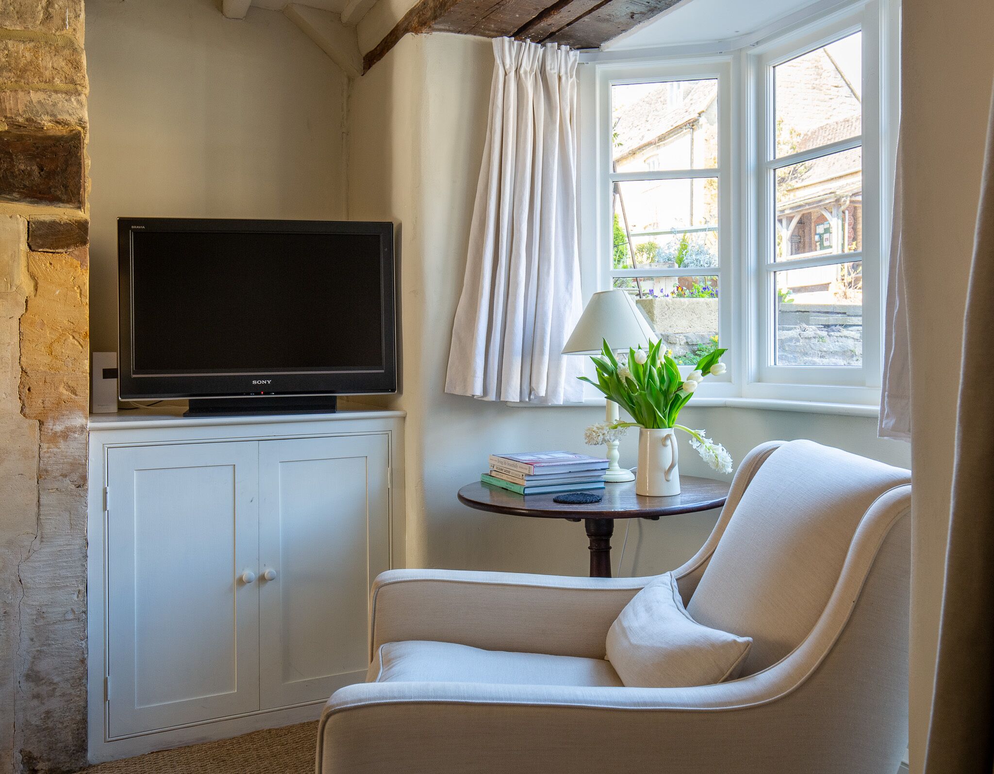 A cozy room with a flat-screen TV on a cabinet, a comfortable chair, a small table with a lamp and a vase of flowers, and a window with curtains.