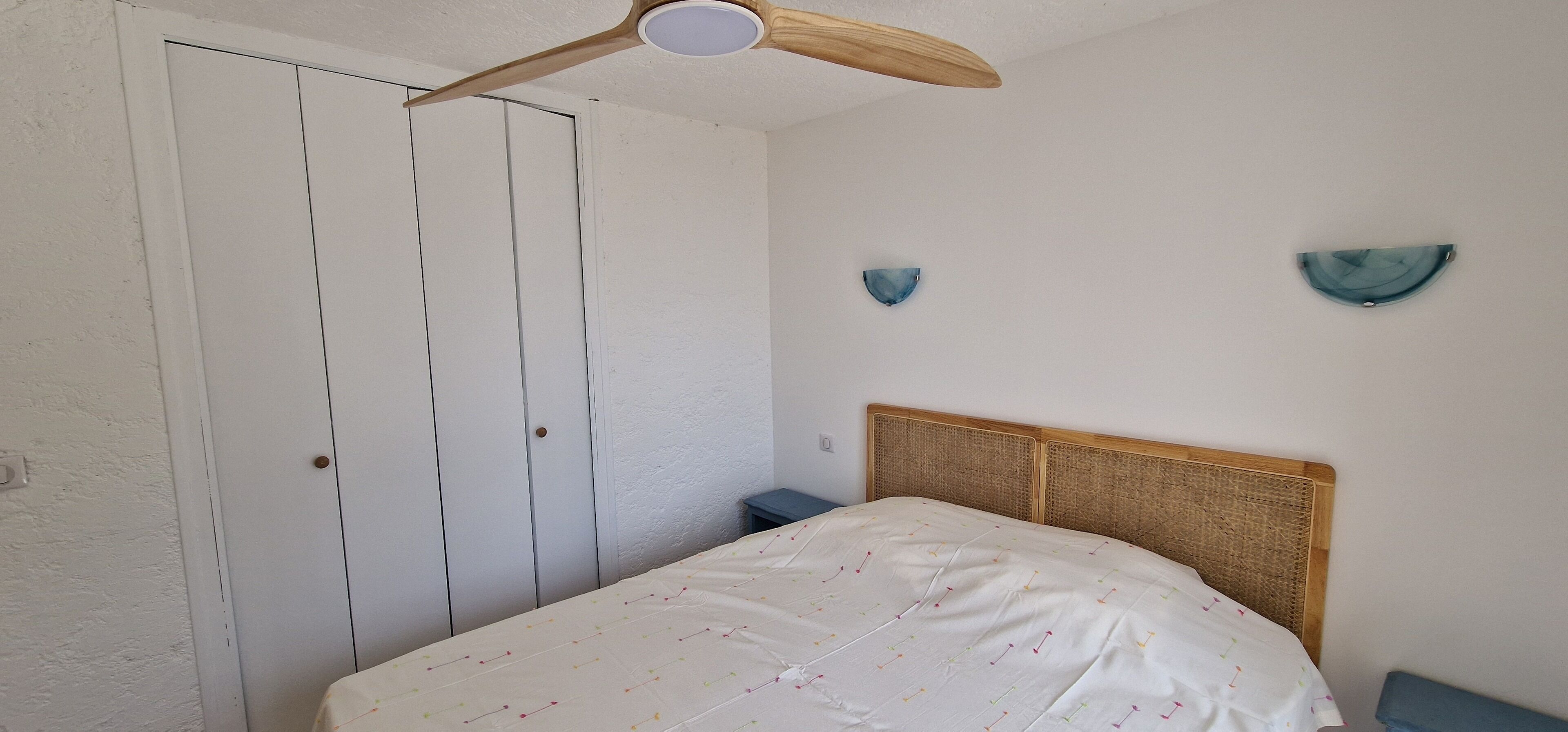 Une chambre à coucher comprenant un lit, un ventilateur de plafond en bois, une armoire et deux appliques murales.